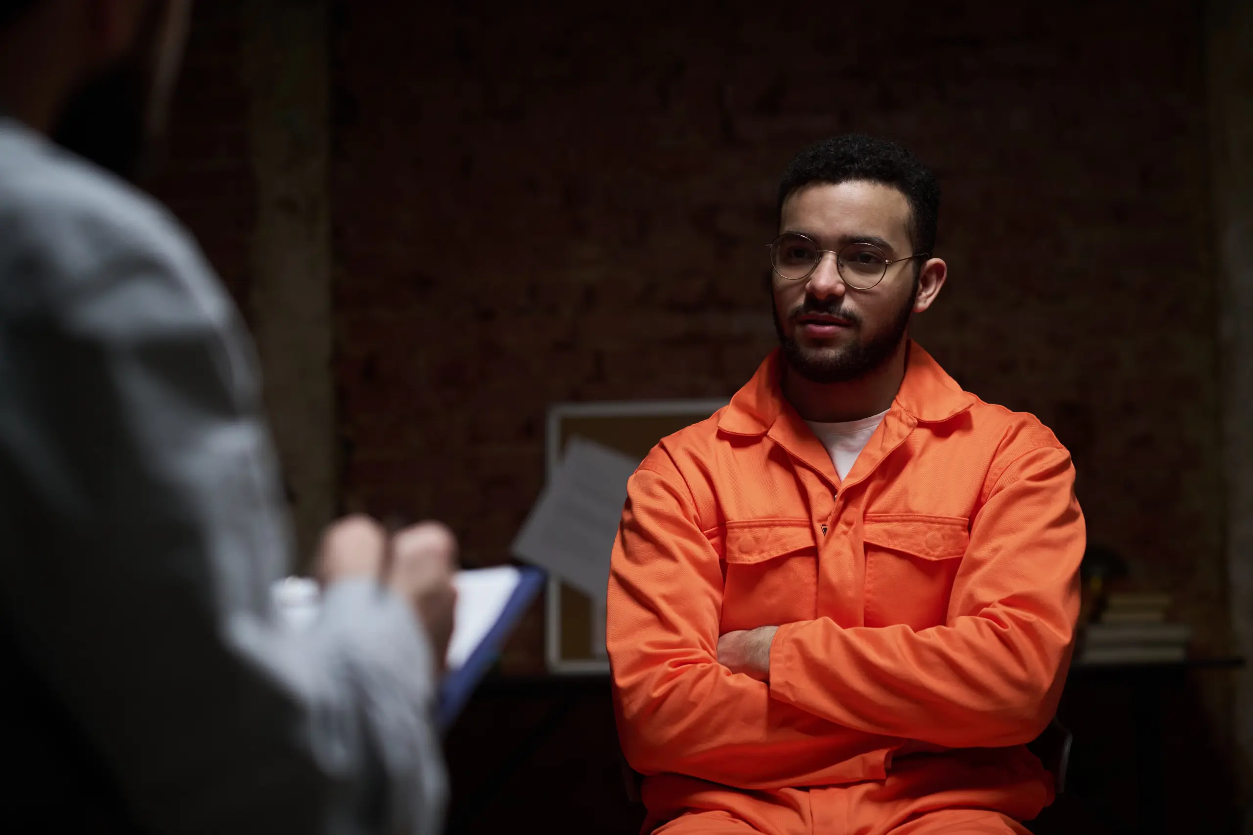 Young adult man sitting with arms crossed wearing prison uniform engaging in group therapy session with unseen counselor holding clipboard in correctional facility setting